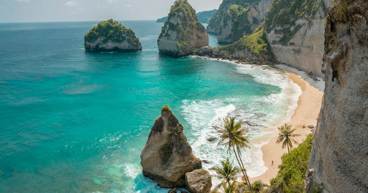 A rock pinnacle in turquoise water and a palm-fringed beach below the cliffs of Nusa Penida’s east coast