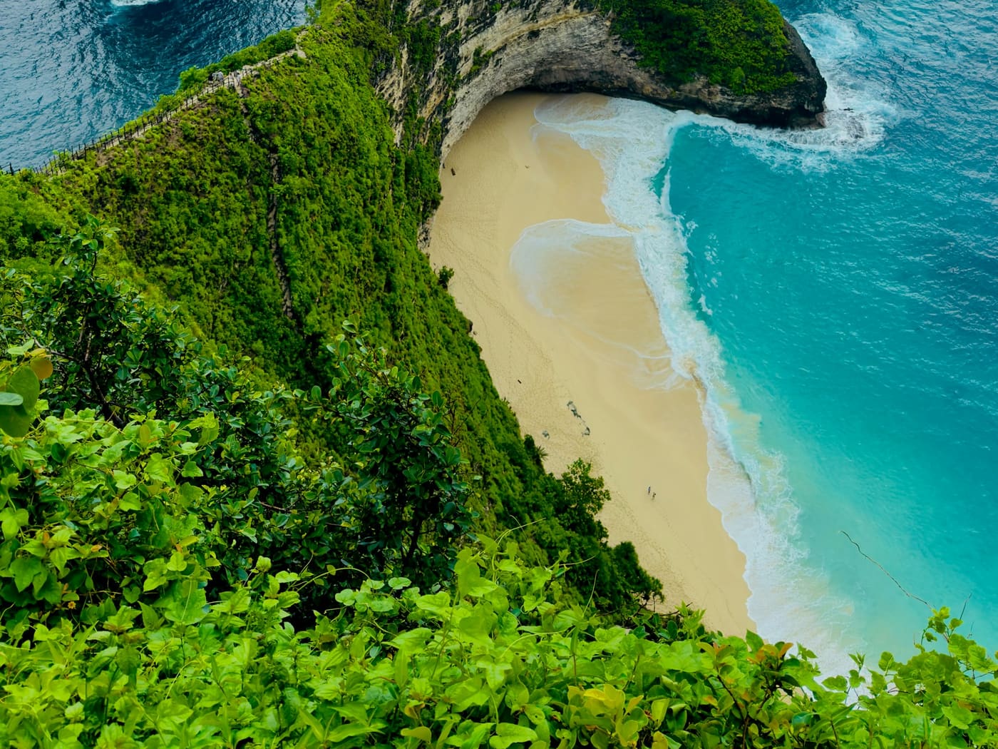 Kelingking Beach from above, with the fenced ridge path running down the green headland towards the sand